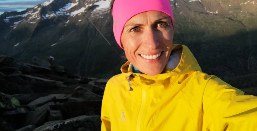 eine Frau mit gelber Jacke und rosa Mütze macht ein Selfie im Freien in den Bergen